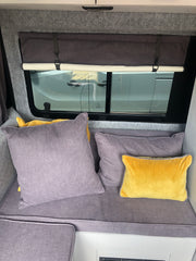 A view of the inside of a recently done up camper van. We can see two bench cushions in a medium grey fabric with a selection of scatter cushions in the same grey fabric and a bright yellow fabric. Behind the cushions we can see one of the van's side windows which has a rolled up black out blind in the same fabric up on it.