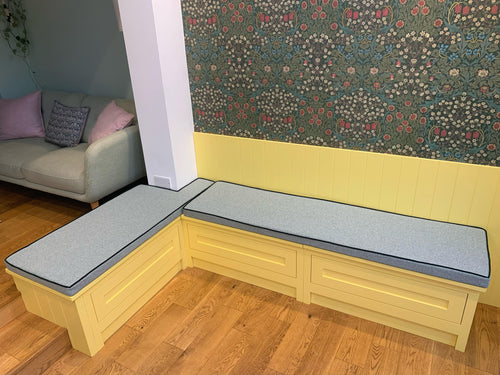 A brightly coloured dining bench area with daffodil yellow panelled walls and a dark green floral wallpaper. The wooden built in bench seating area has two custom shaped bench cushions on top. They have firm foam filling and are covered in a flecked light green upholstery fabric. The cushions have dark green contrast piping and have concealed zips.