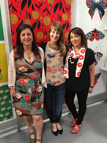Allison Sloan Polish and Tara Reed of Spoonflower visit Misha Zadeh in her booth at Surtex 2017 in NYC. Misha's Rain Garden in Tomato Wall paper sits prominently behind them. All three women are wearing clothing with prints from Spoonflower. Misha is wearing her Inky Poppies print as a scarf.