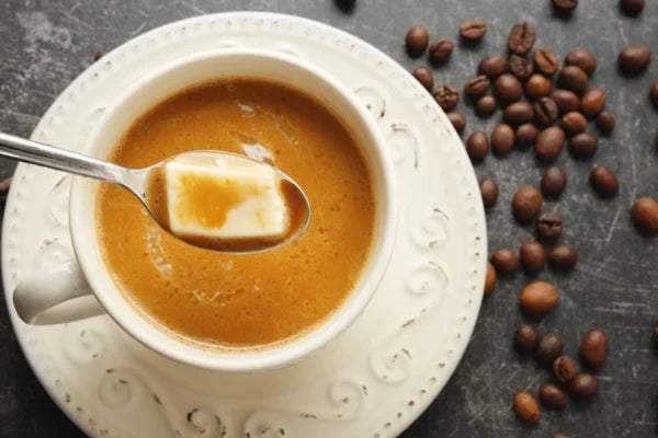 A cup of coffee with a spoon and creamy butter cube on a decorative saucer, surrounded by scattered coffee beans.