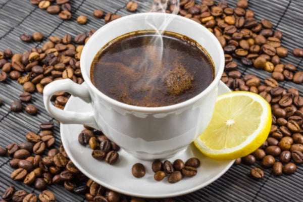 A cup of hot coffee with a lemon slice on the side, surrounded by coffee beans on a textured surface.