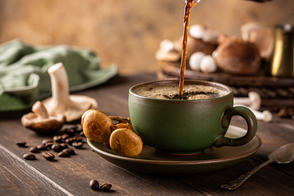 Coffee being poured into a green cup surrounded by coffee beans and mushrooms.