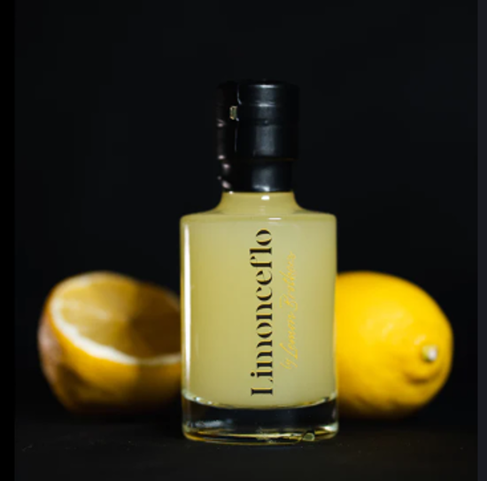 A miniature bottle of Lemon Brothers’ Limonceflo with two lemons positioned behind it, set against a dark background.