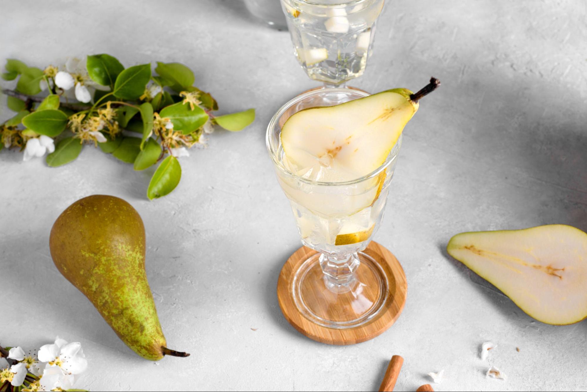 Limoncello and pear cocktail in a flute, garnished with a pear slice, with the fruit, leaves and blossom from a pear tree on a white table.