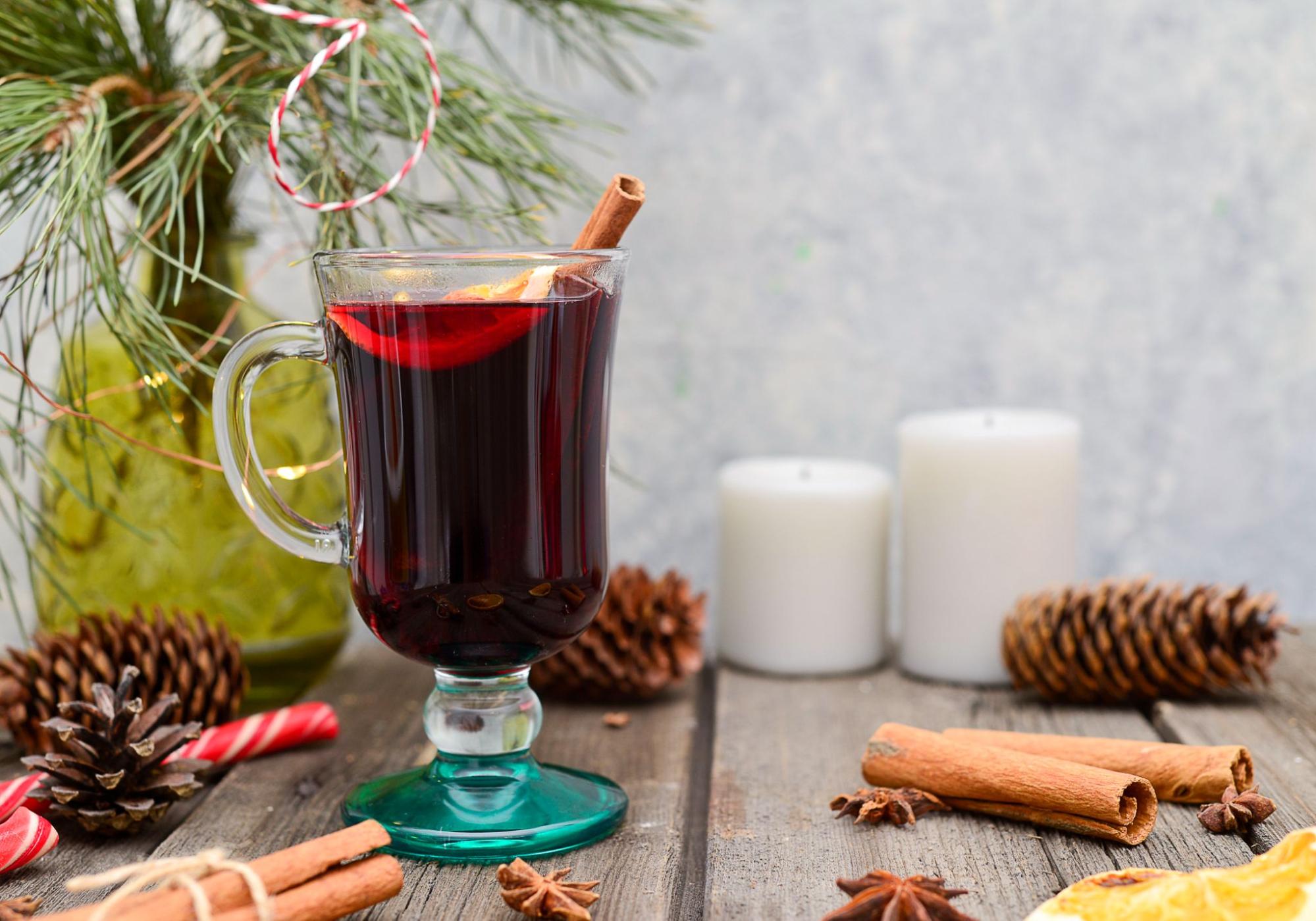 Limoncello Mulled Wine in a glass latte mug, garnished with a lemon slice and cinnamon surrounded by spices, pinecones, two candles, and foliage.