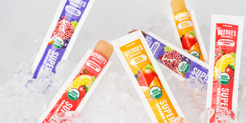 An image of six DeeBee’s Organics Classic SuperFruit Freezie Pops standing atop a bed of ice. Two of the refreshing freezie pops are open, and ready to eat.