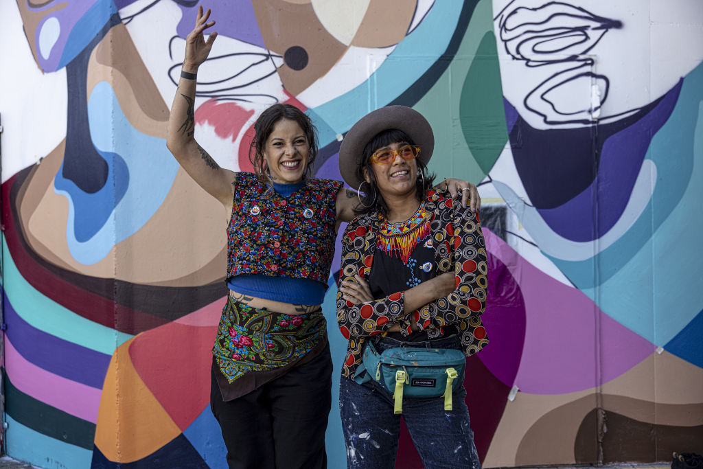 The artists Jumu Monster and Mari Pavanelli stand in front of their mural hugging and laughing towards the camera.