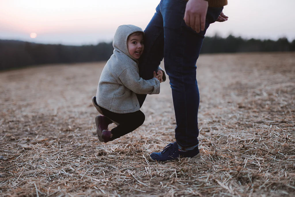 Evening mood, a field with straw, in the picture the legs of an adult person, lifting one of their feet a bit with a little child sitting on it, holding on to the leg with both hands and looking exuberant.