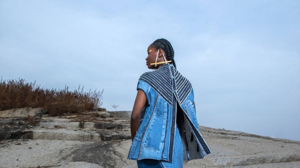 Back and profile of a person in clothes of the label NKWO. The person is standing on a rocky ground running towards a wide, slightly cloudy sky.
