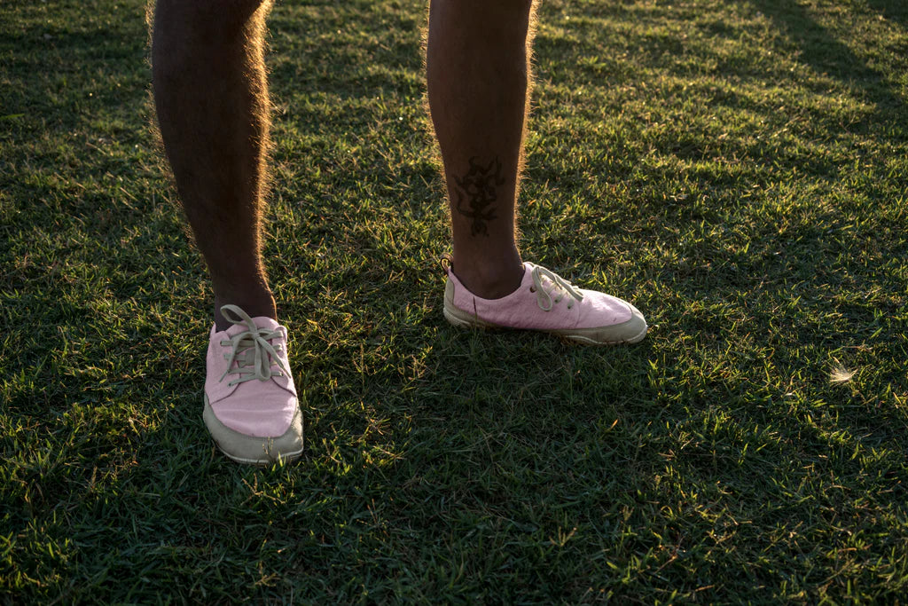 Lower leg of a person, bare hairy legs and a black tattoo above their ankle. The person is wearing pink Wildling Shoes minimal shoes and is standing on a mowed meadow.