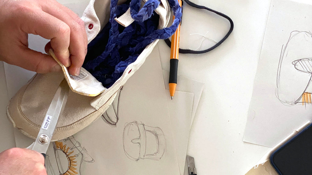 Close-up of a Wildling Minimal shoe on a table being worked on by two hands with scissors and ribbons. On the table next to it are sketches of a new shoe model.