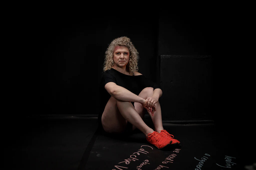 A person with blond curls, black dress, bare legs, and red wildlings is sitting on the ground and looking seriously into the camera. Ground and background are black.