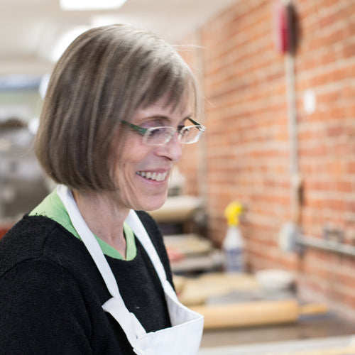 Woman with glasses and an apron looking down and smiling against brick background