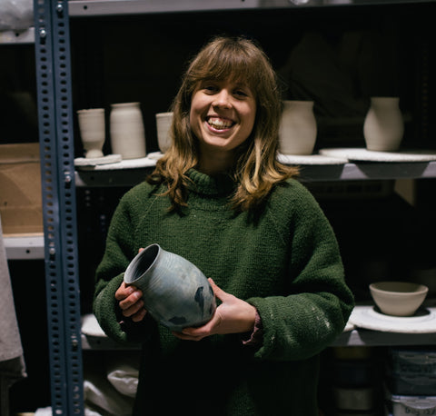 Kate Welton ceramicist, a young woman in a green jumper holding a ceramic pot in front of shelves filled with other ceramic pieces 