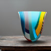 a tall and gently curved multi-coloured glass vase on a wooden table by Ruth Shelley 