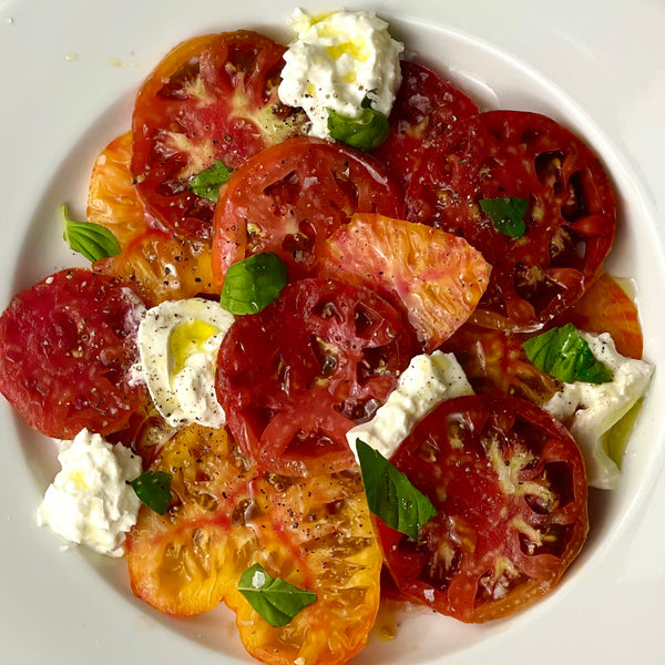 Juicy slices of heirloom tomatoes of various colors, sizes, and shapes are artfully arranged in a deep round plate. Spoons of creamy burrata and torn basil leaves adorn the whole, which is drizzled generously with EVOO.