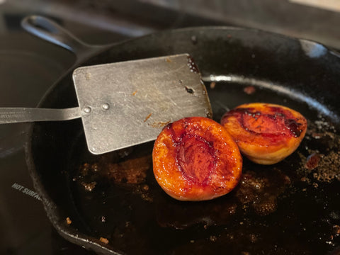 Two peach halves glistening in dinner's pan drippings while being roasted in a 12" cast iron skillet