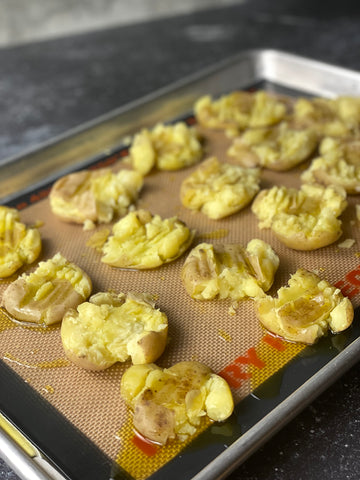 A baking sheet with Silpat 16 small Yukon Gold potatoes, smashed, covered in oil, and waiting to be put in the oven for finishing.