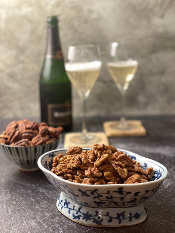 half a pound of fire dried walnuts is in the foreground in a Chinese pedestal dish, a small round dish of pecans beyond it, and in the background, a bottle of sparkling wine and two filled glasses