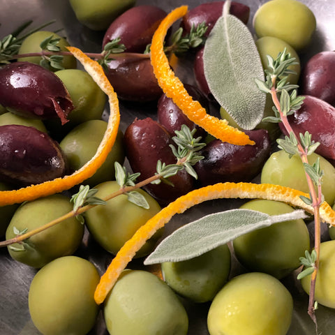 close up image of large round and medium olive olives with sage, thyme, rosemary, and strips of orange peel