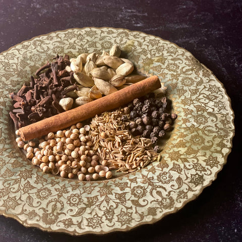 A heavily figured brass and enamel plate with delicately scalloped edges holds fresh whole cloves, cardamom pods, a cinnamon stock, coriander, cumin, and tellicherry black peppercorns