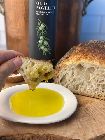 Liz dips a torn piece of Dan's country loaf into a plate of Olio Novello. Salt, oil, and bread are in the background.