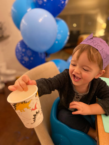 Freddie smiling at his dinosaur party cup with blue balloons in the background