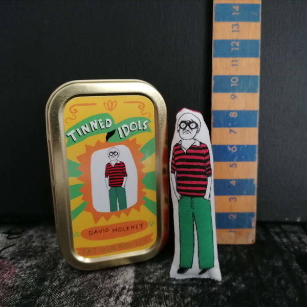 Mini fabric doll of artist David Hockney beside a gold tin and a ruler for scale against a black backdrop.