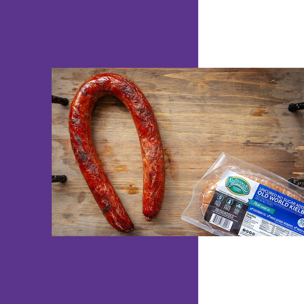 An overhead image of a grilled rope sausage sitting on a wooden cutting board with a pack of Pederson's Farms Uncured No Sugar Added Old World Kielbasa sitting next to it.