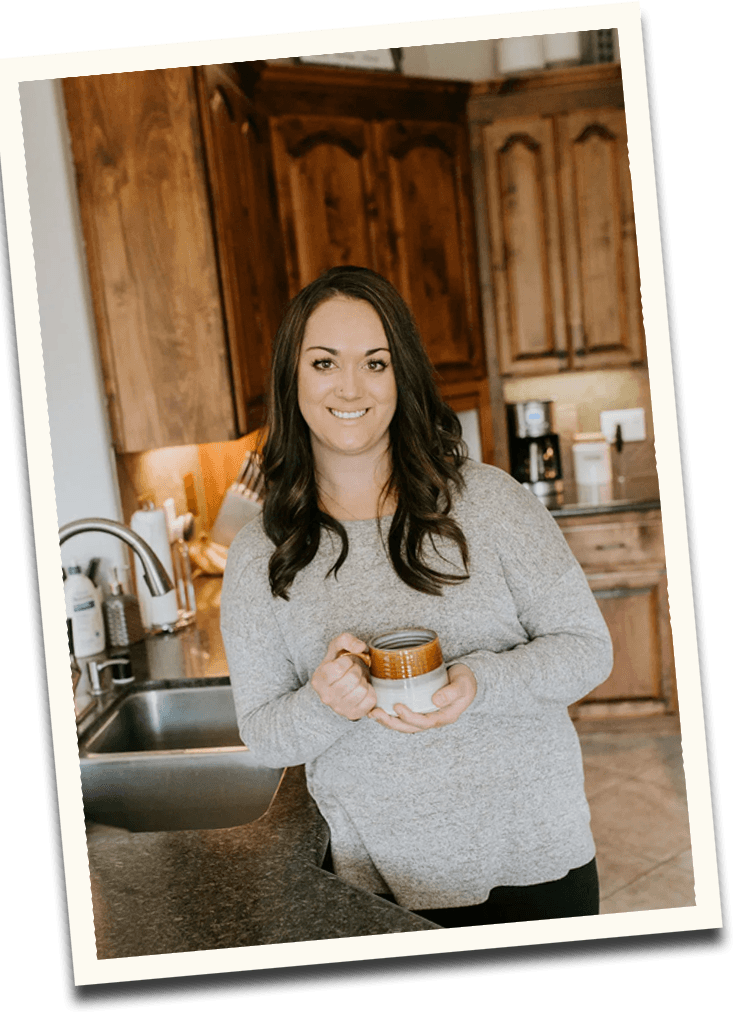 A photo of Emilee Mason, standing in a kitchen, leaning against the counter. Emilee is wearing a grey blouse and holding a brown and white coffee mug!