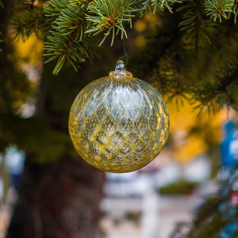 An ornament, hanging from an evergreen tree in front of a blurred winter background. The ornament is whiskey colored and utilizes the pineapple mold, adding fractal reflections of the world around it.