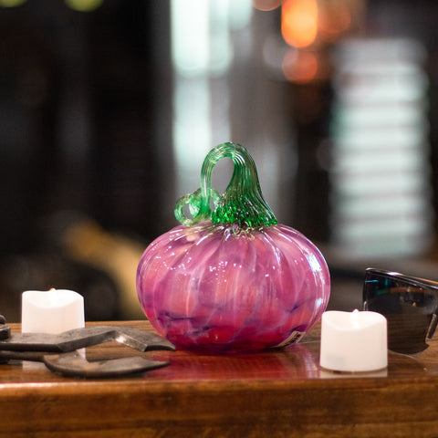 A pink glassblown pumpkin with a green stem sitting on a counter, surrounded by glassworking tools and electric tealight candles.