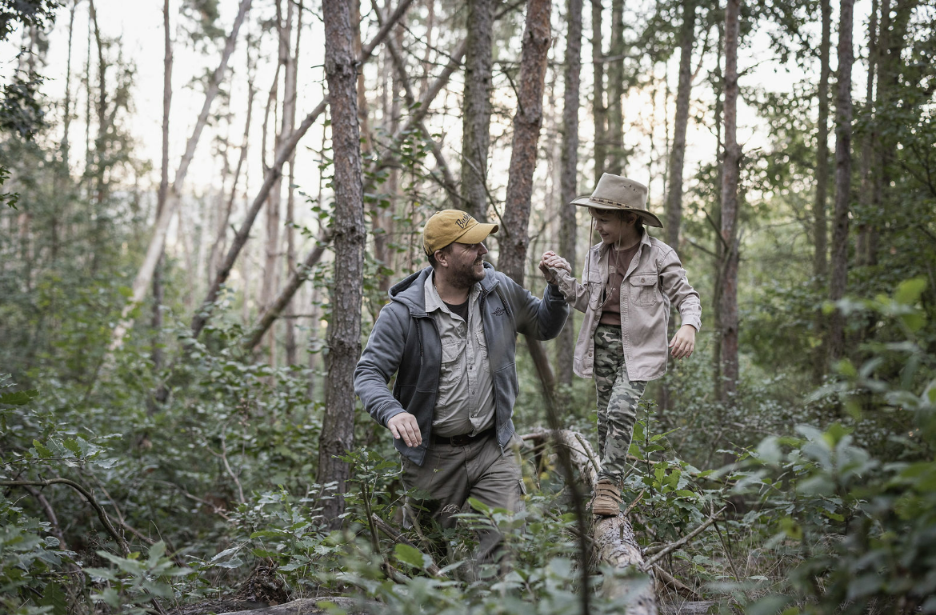Odrasel moški drži majhnega otroka, ki hodi po lesenem deblu v gozdu in se smeje s klobuki na glavi | BUSHMAN
