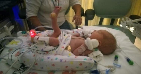Bee laying in a NICU pod, he has a draining tube in his mouth, and white mittens on his hands, he also has one foot sticking up in the air