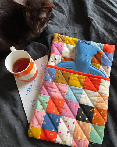 quilted hot water bottle cover on a bed with a cat, book and cup of tea