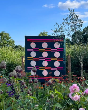 a dark gree quilt with pale pink circles and multi-coloured stripes displayed in a flower garden
