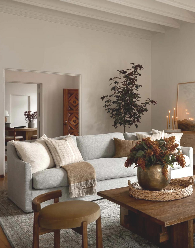 Be patient, creating a collected home style. Scene of living room with light colored sofa, cozy neutral throw pillows, accent stool, and styled coffee table with vase and woven tray.