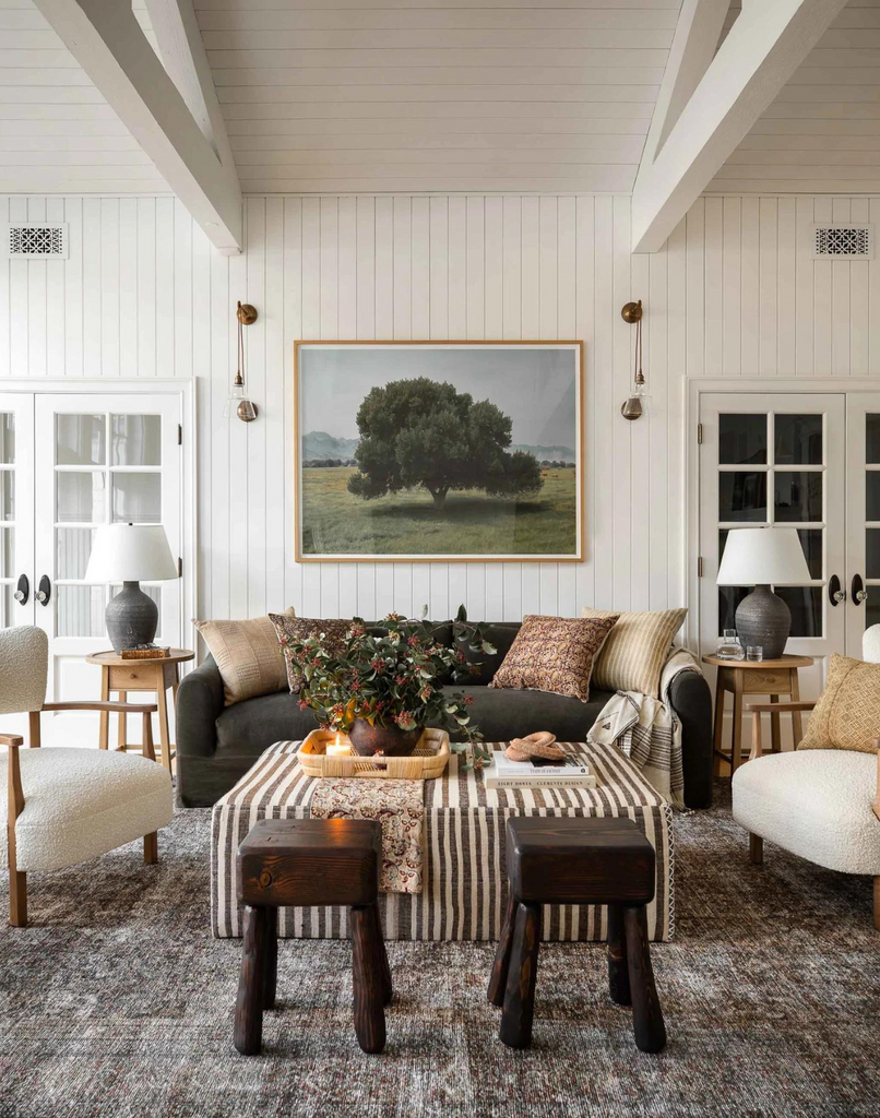 Shop a variety of stores to create a collected home style. Living room scene with dark sofa, boucle accent chairs, wooden stools, landscape artwork, and styled coffee table with decor.