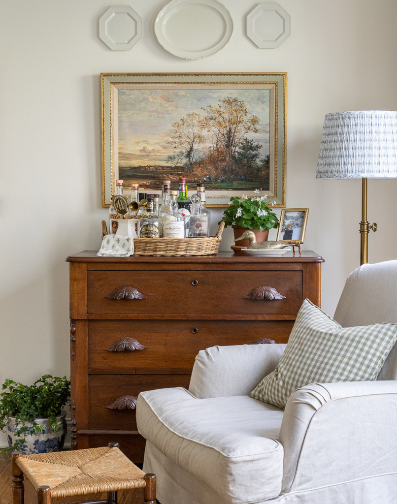 Think outside the box with decor to create a collected home style. Living room vignette scene of vintage dresser being used as a bar cart. Basket tray with liquors, vintage art, and vintage dishes hung on the wall as art.