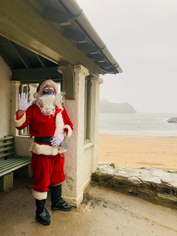 Father Christmas standing under the shelter at Readymoney Cove, Fowey. He is waving. The sea is in the background.