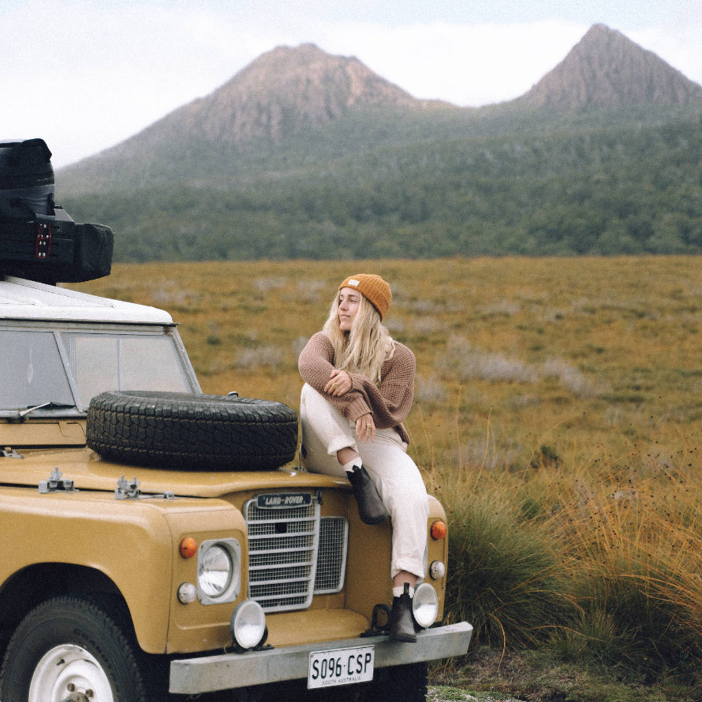 Woman with long blonde hair wearing a beanie sitting on a 4wd in mountain valley