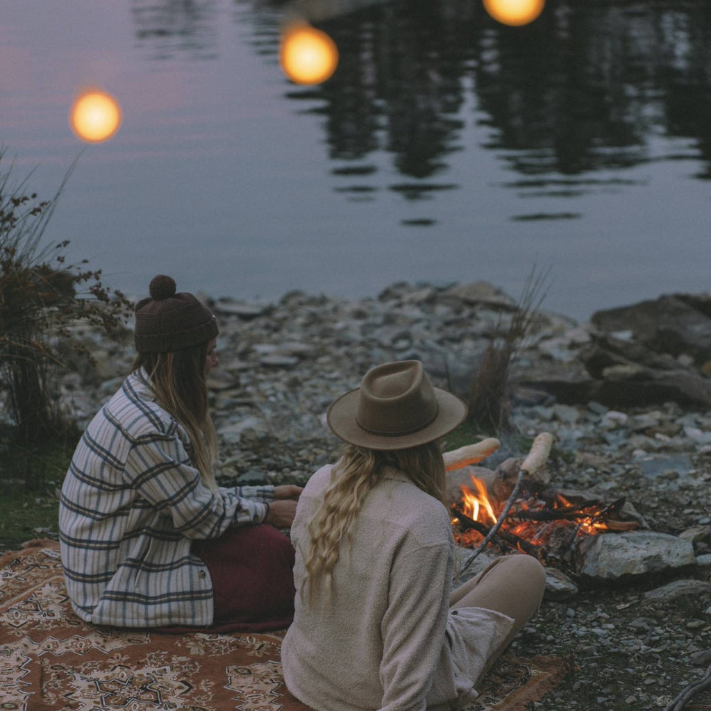 Two women, one wearing a beanie and one wearing a wide brim wool hat are sitting by a camp fire