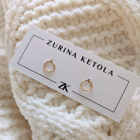 Zurina Ketola's circle post earrings in 14K gold fill on an earring card, perched on top of a cozy white knit blanket.