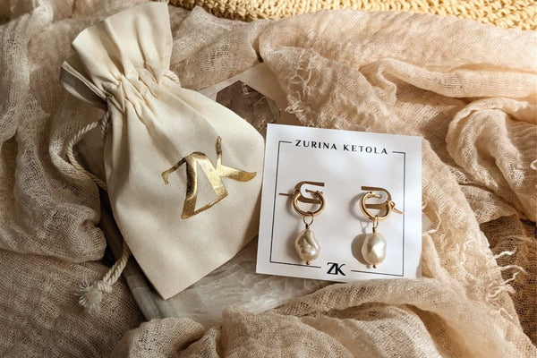 This image shows a pair of small gold hoops with luminous white pearls that have an organically imperfect shape. The earrings are on an earring card from the designer Zurina Ketola, and are laying on top of a summery beige fabric, and sit next to a beautiful drawstring cotton pouch with a golden ZK logo. 