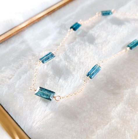 Zoomed in side view of the moss aquamarine necklace, featuring a pattern of moss aquamarine baguettes connecting delicate 14K gold fill chain. The image only shows the bottom of the necklace, which is laying flat in a glass jewelry box with brass edge detail on a white background in the sunlight.
