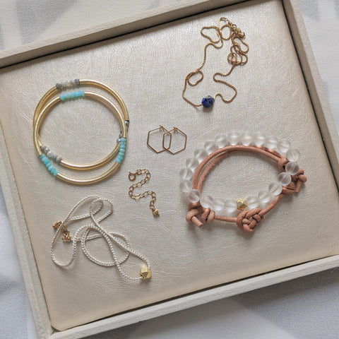 An overview of a selection of gold jewelry in a white tray lined with pearlized vegan leather. One pair of hoops, one necklace extender, two necklaces and four bracelets. 