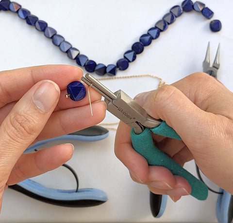 Overhead view of jeweler's hands using pliers to to make a lapis pendant for a gold necklace.