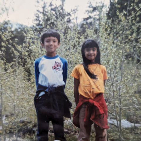 This is an image of a brother and sister in the woods. Both children have olive tone skin and black hair. The boy has straight short hair, and the girl has bangs and long hair. They're likely on a hike or camping, and smiling at the camera.