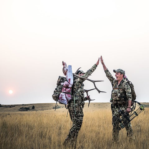 Sitka Gear - two men high fiving at dawn with one of the two men having a recently harvested Elk by compound bow on his back