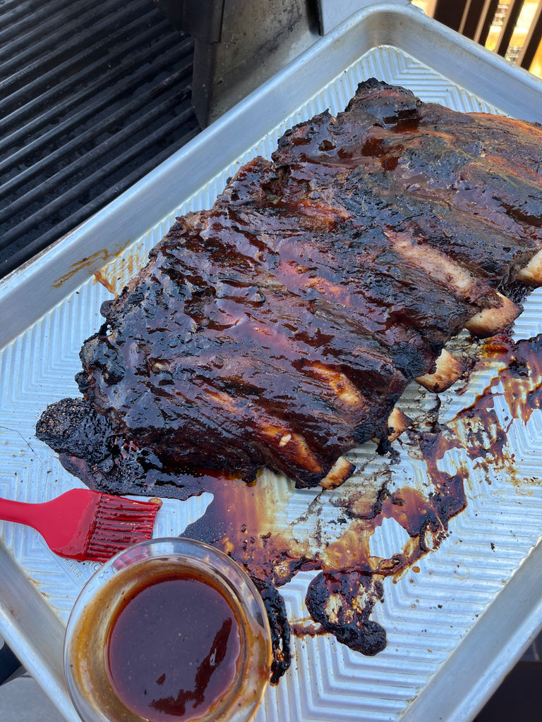 Barbecue bison ribs on a pan next to a grill, alongside a brush with barbecue sauce